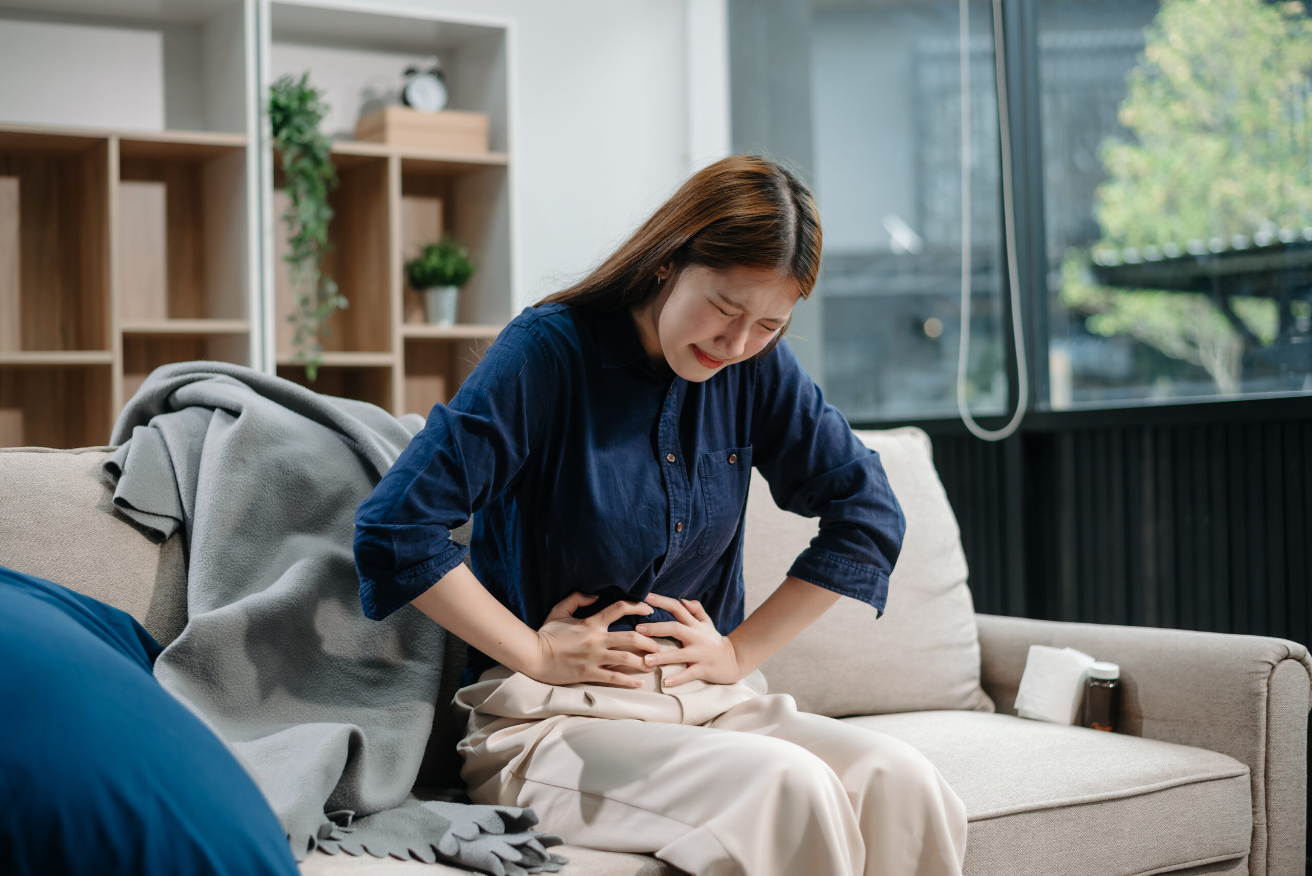 Young Asian woman suffering from flu symptoms, covered with a blanket on a sofa. Concept of illness, healthcare, and recovery. Stay indoors, stay healthy