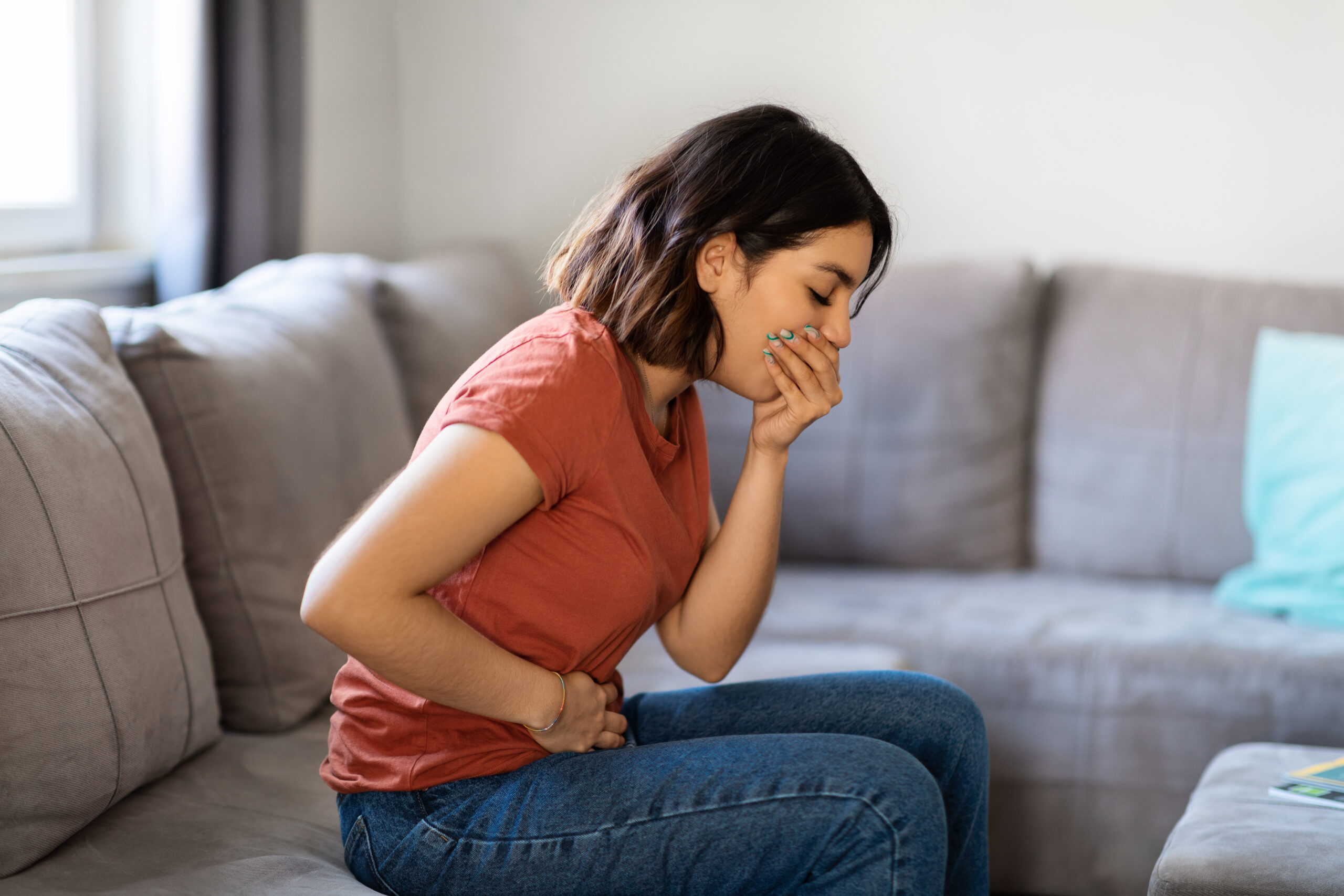 Food Poisoning Concept. Young Arab Female Feeling Sick At Home, Ill Middle Eastern Woman Suffering From Nausea, Covering Mouth And Touching Belly While Sitting On Couch In Living Room, Side View